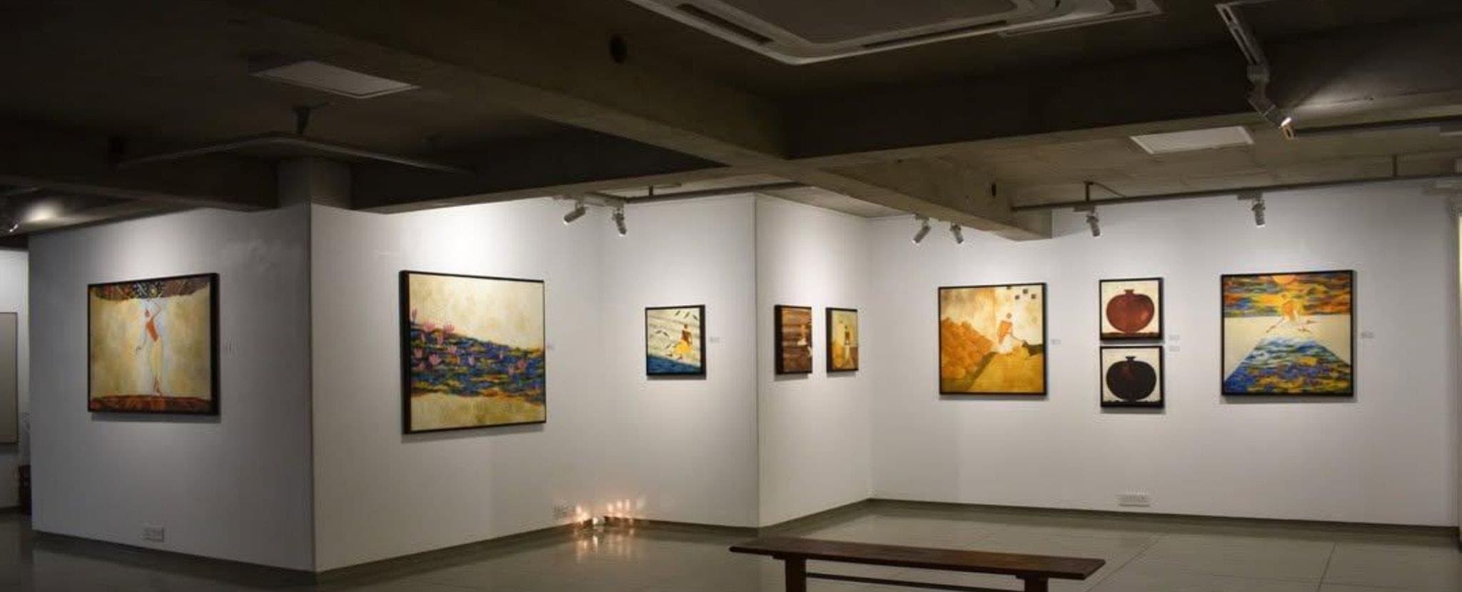 A dimly lit gallery with paintings on white walls, a wooden bench and small lamps on the floor.